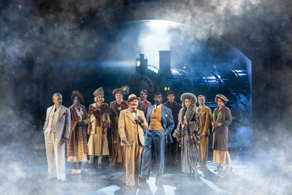 image of cast standing together in front of a train situated in smoke in the background