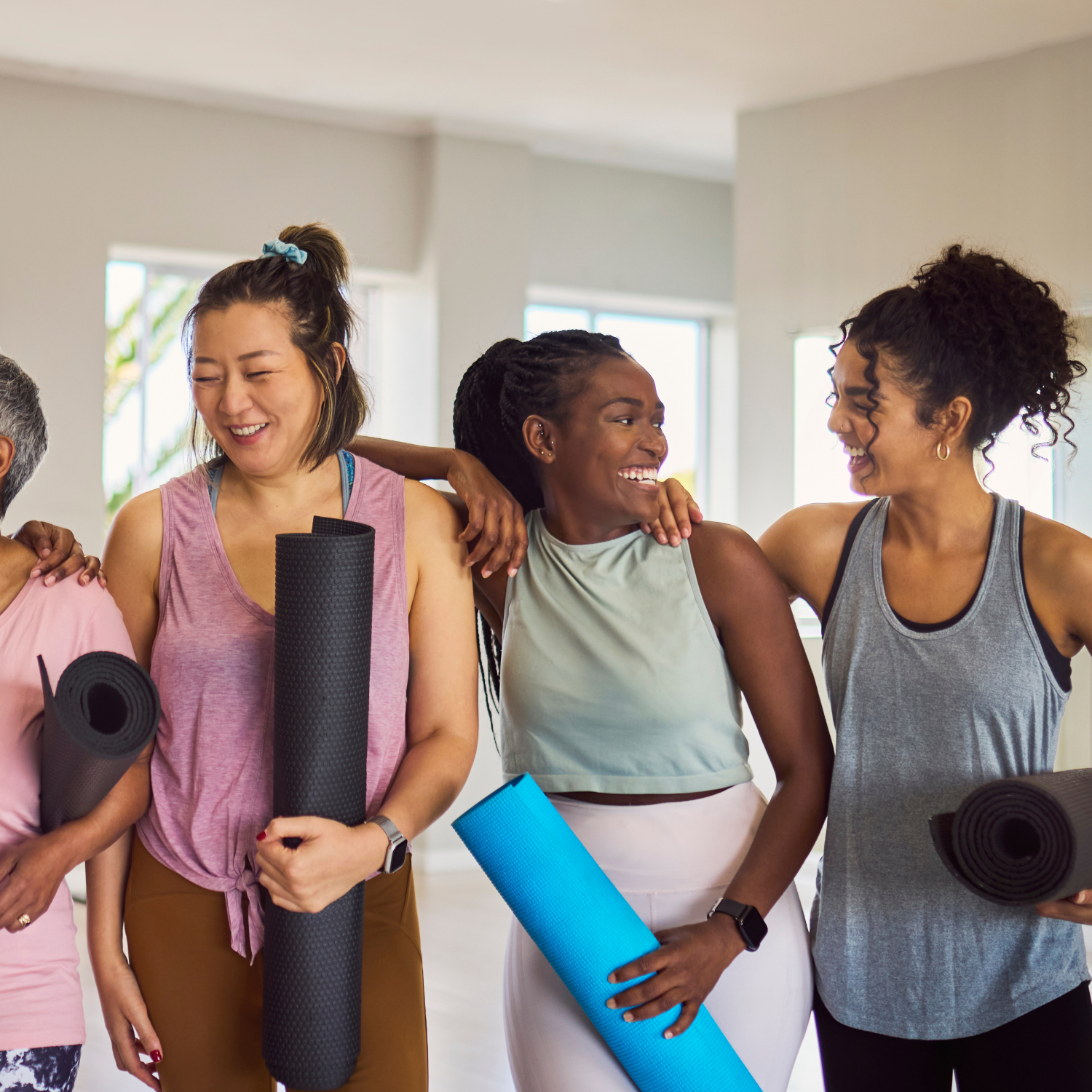 Group of diverse female friends in yoga attire, standing in a row with yoga mats, laughing excitedly and looking at each other before exercise in studio
