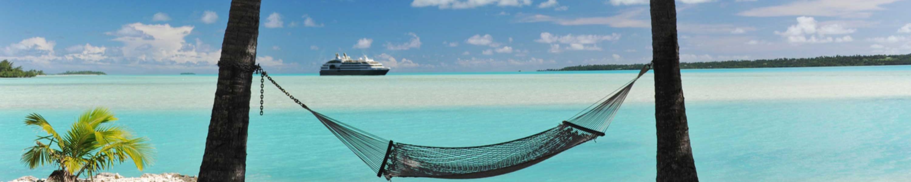 A cruise ship appears on the horizon of a tropical turquoise lagoon - with a hammock in semi silhouette, shaded by palm trees