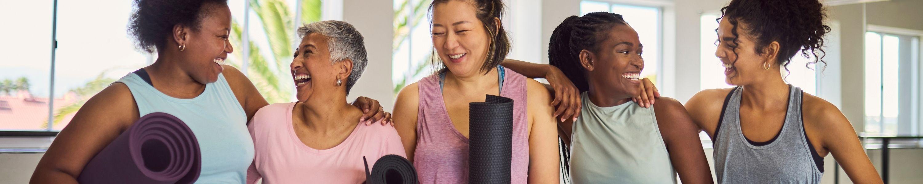 Group of diverse female friends in yoga attire, standing in a row with yoga mats, laughing excitedly and looking at each other before exercise in studio