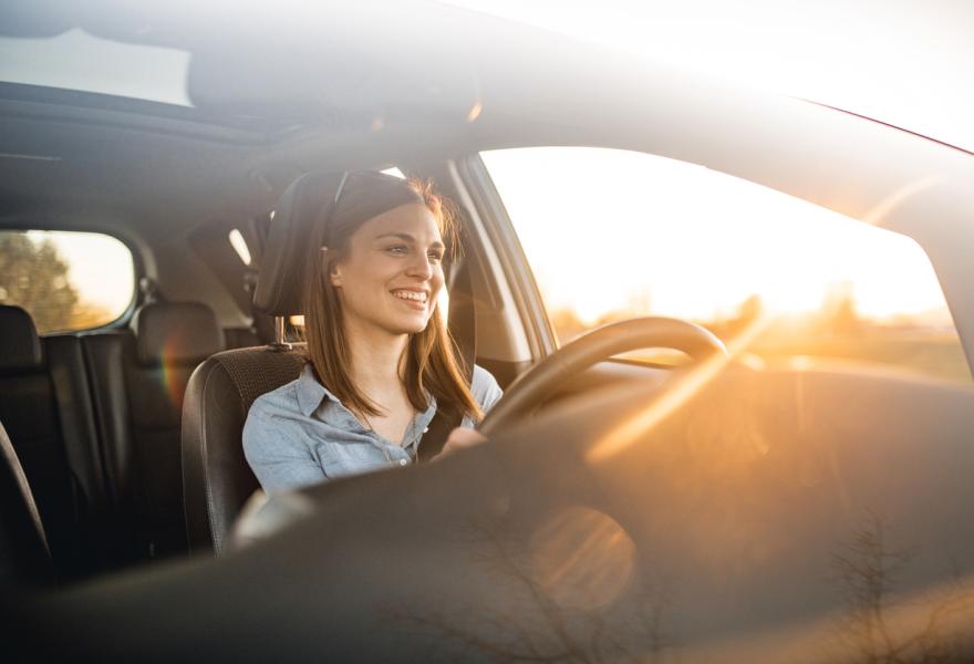 Young woman in a car. She is driving, smiling and looking out. Sun is shining from back on a sunny spring afternoon.