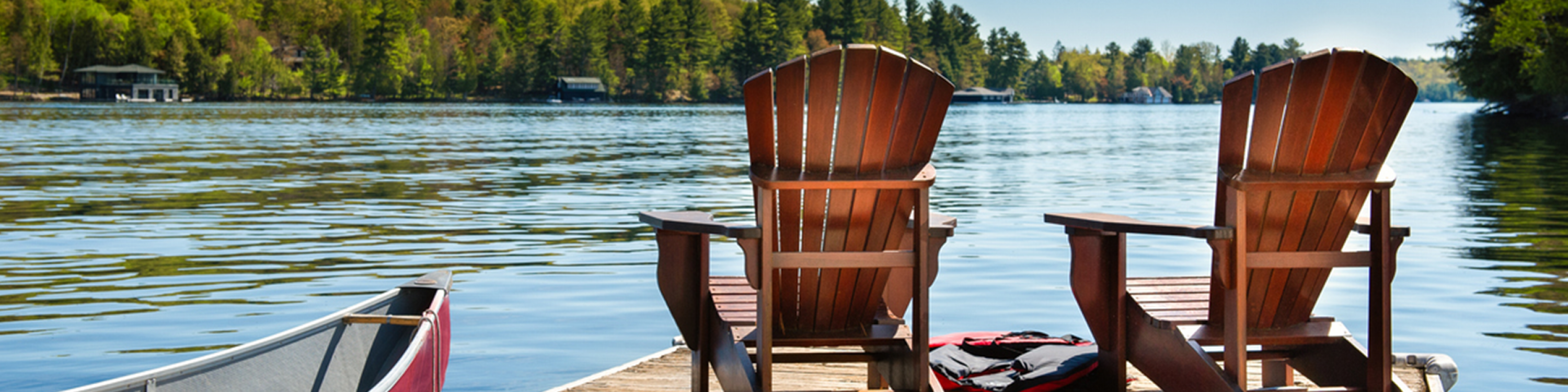 Two Muskoka chairs on a wooden dock overlooking the blue water of a lake in Muskoka, Ontario Canada. A red canoe is tied to the pier and life jackets are visible near the chairs.